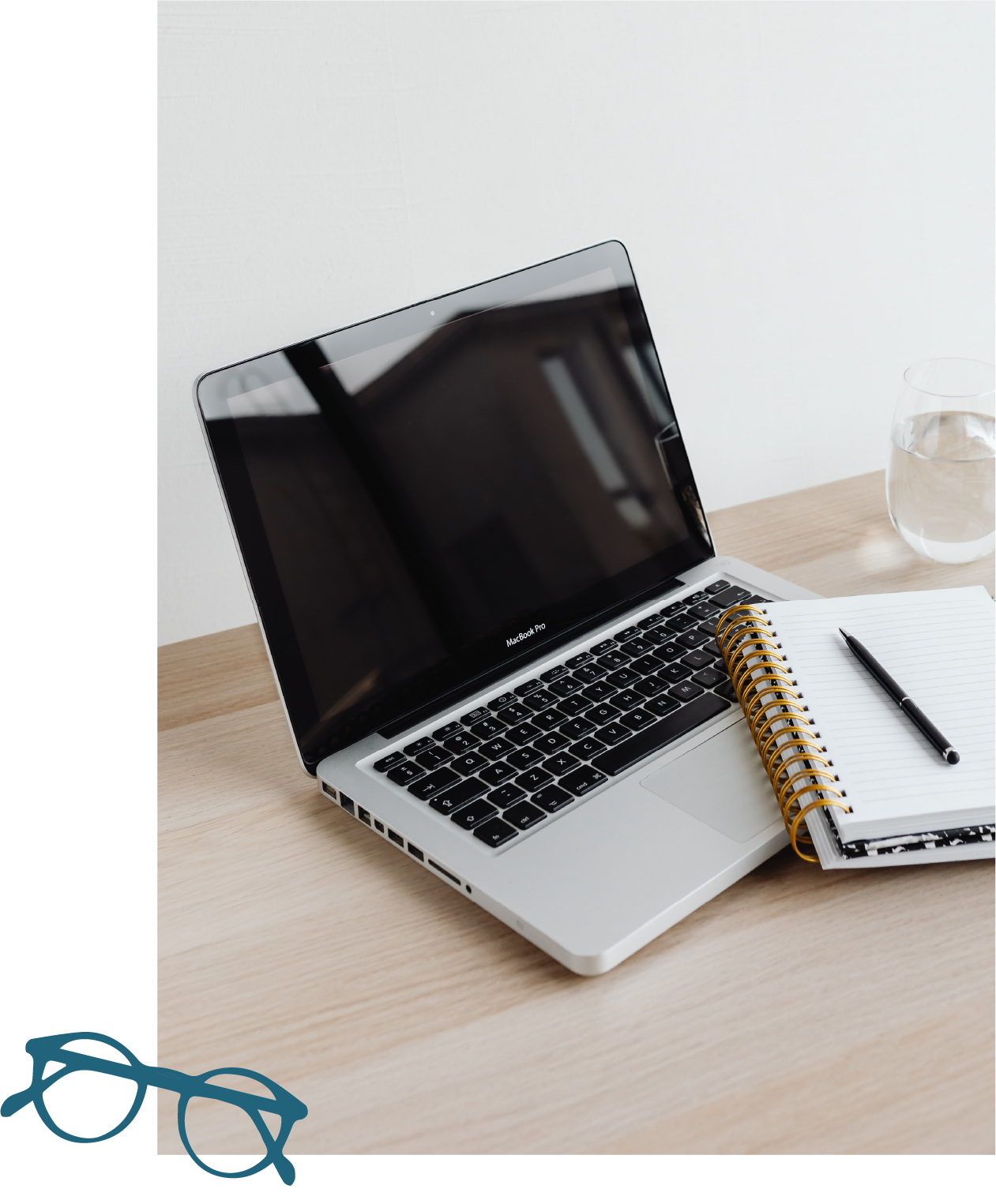 Asset 4@3x ScopeSchool A laptop on a wooden desk next to a spiral notebook with a pen, a glass of water, and an illustrated pair of glasses in the bottom left corner.